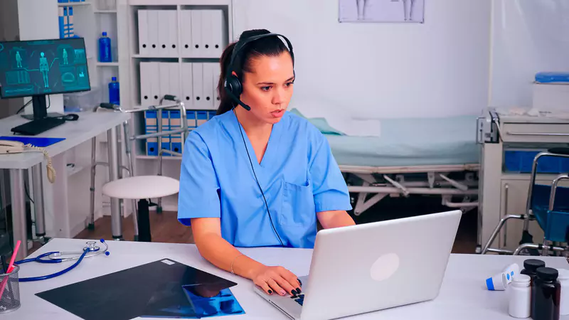 Informatics nurse using headset and laptop, managing clinical data to optimize healthcare workflows