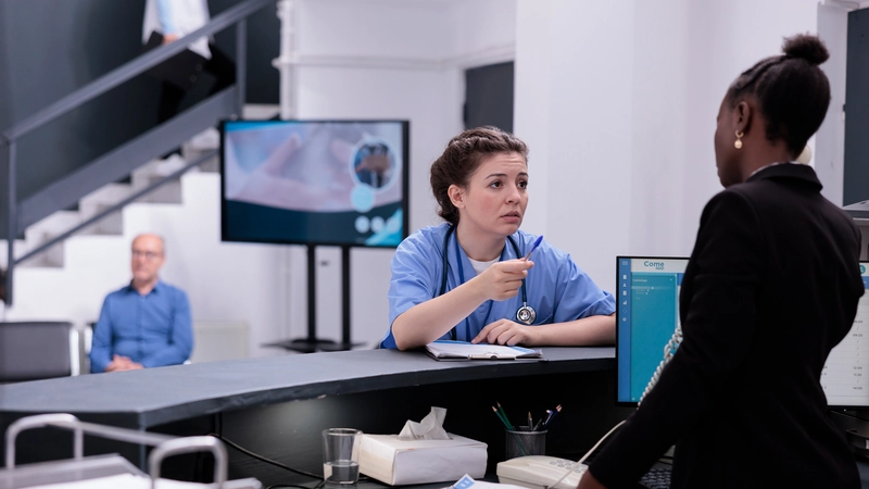 Nurse in blue scrubs speaking with a colleague at the hospital reception desk, showing signs of stress and workplace tension