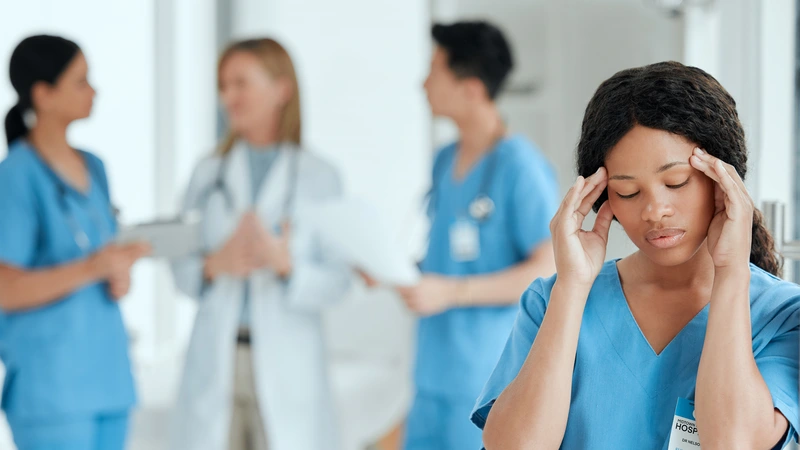 Stressed nurse holding her head in frustration while colleagues talk in the background, representing the emotional toll of hospital politics