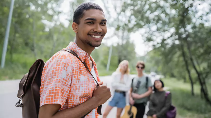 young-smiling-student-with-backpack-standing-outdoors-with-group-in-background